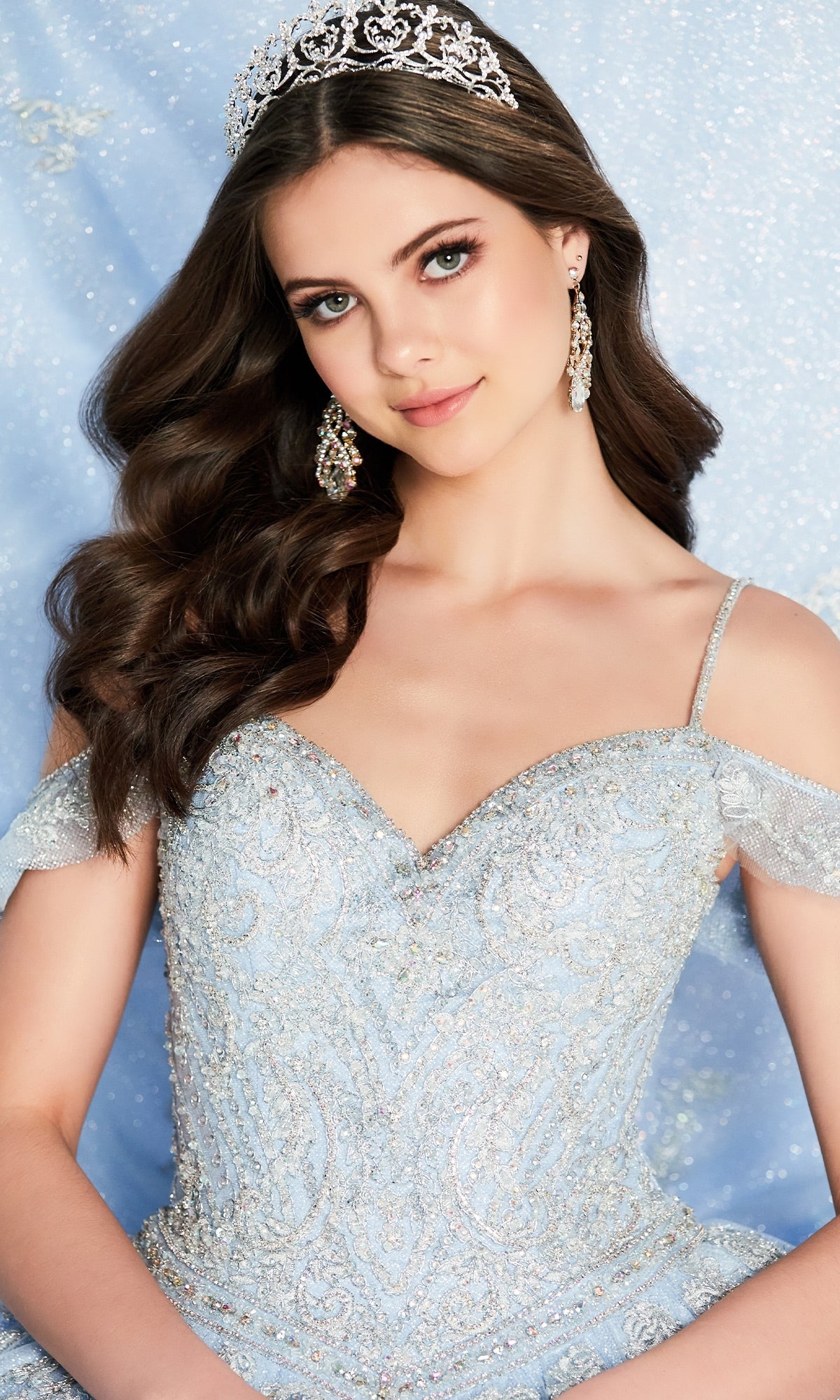 A young woman in the Light Blue Quinceanera Dress: Princesa PR22032, featuring an off-shoulder design and lace appliques, poses with a sparkling silver tiara against a shimmery blue background.